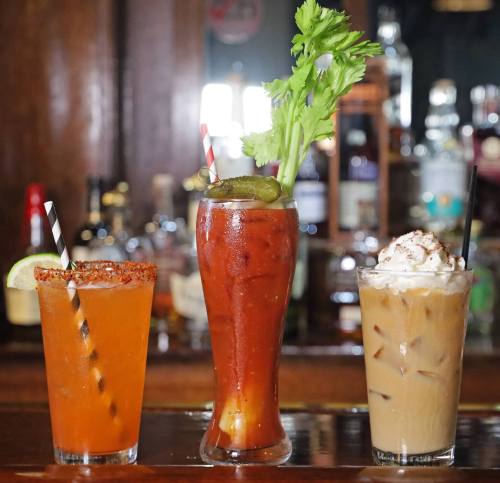 Three colorful cocktails on a bar: a spicy drink with a celery garnish, a fruity orange drink, and a creamy iced coffee.