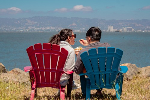 A couple sits in colorful chairs by the water, enjoying a meal with a scenic view of the city in the background.