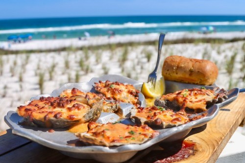 A platter of baked oysters topped with cheese, served with a lemon wedge and a bread roll, with a beach backdrop.