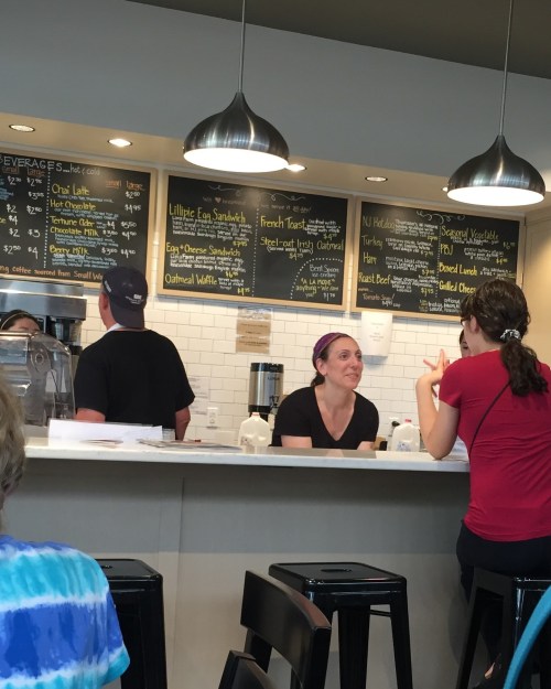 A café scene with a counter, menu board, and two people chatting; one is a barista, the other a customer.