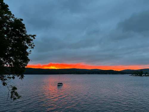 A serene lake at sunset, with vibrant orange and red hues in the sky and a lone sailboat on the water.