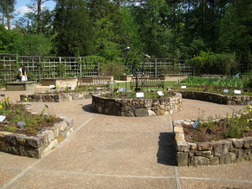 A landscaped garden with stone beds, labeled plants, and a central circular area surrounded by greenery and benches.