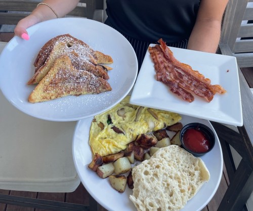 A person holds two plates of breakfast: French toast with powdered sugar, bacon, and an omelet with potatoes and bread.