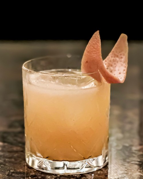A refreshing cocktail in a glass, garnished with a pink citrus peel, resting on a dark countertop.