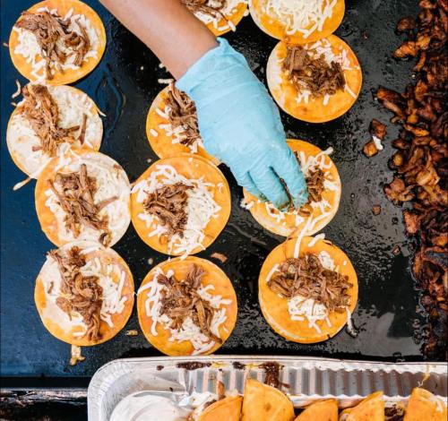 A hand in a blue glove adds shredded meat and cheese to corn tortillas on a hot grill.