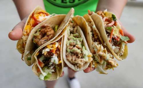 A person holds four colorful tacos filled with shrimp, chicken, and various toppings, showcasing a delicious meal.
