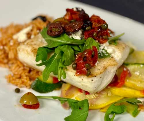 A plate featuring grilled fish topped with olives, capers, and arugula, served with rice and sautéed vegetables.