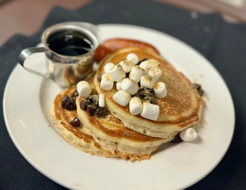 A plate of fluffy pancakes topped with chocolate chips and mini marshmallows, served with syrup and a side of sauce.