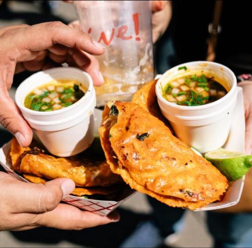 Two hands hold cups of soup and a tray with two golden, crispy tacos, garnished with lime and herbs.