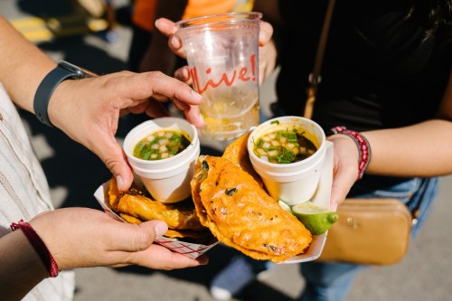 Two hands hold a plate of golden empanadas and two cups of soup, with a drink in the background.