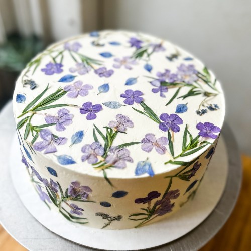 A round cake decorated with edible flowers and green leaves on a white frosting background.