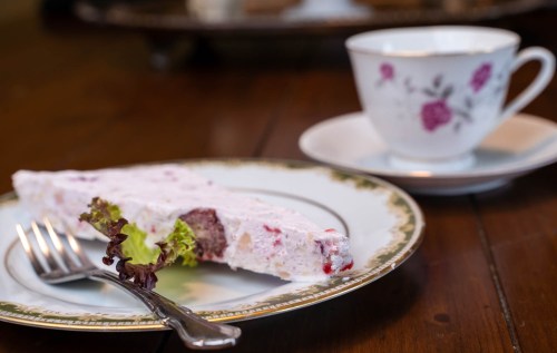 A slice of layered dessert on a decorative plate, with a fork and a floral teacup in the background.
