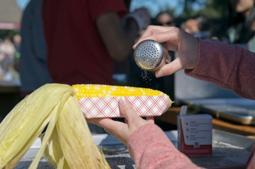 A hand sprinkles salt over a freshly cooked ear of corn on a paper tray at an outdoor event.
