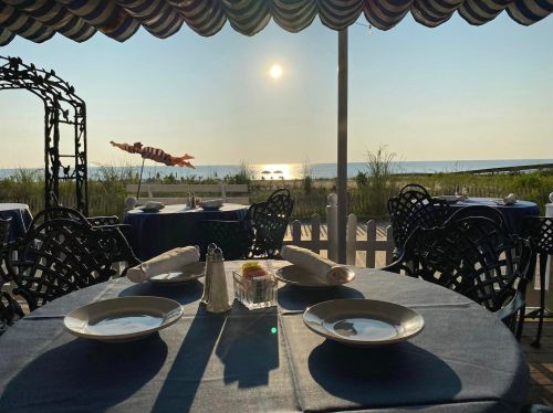 A seaside restaurant table set for dining, with a view of the ocean and sunset in the background.