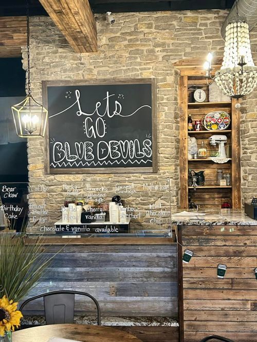 A cozy café interior featuring a chalkboard with "Let's Go Blue Devils" and a wooden counter with various treats.