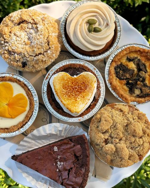 A variety of baked goods on a plate, including muffins, tarts, and a chocolate brownie, garnished with flowers.