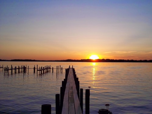 A serene sunset over a calm lake, with a wooden pier extending into the water and silhouettes of distant trees.