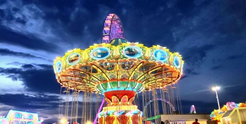 A brightly lit carnival ride against a dramatic evening sky, with colorful lights and a ferris wheel in the background.