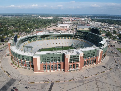 Aerial view of a large stadium surrounded by cityscape and greenery under a clear blue sky.