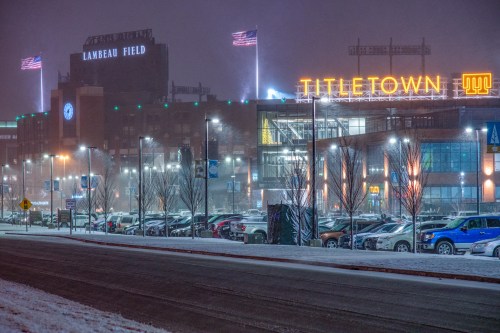 Lambeau Field and Titletown illuminated in a snowy night, with flags waving and cars parked nearby.