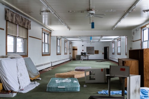 An empty, abandoned room with scattered mattresses, a fan, and a television in the background. Dusty and worn interior.