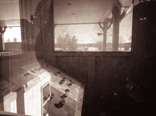 A vintage control room with a panel of buttons, viewed through a window, surrounded by trees and a distant landscape.