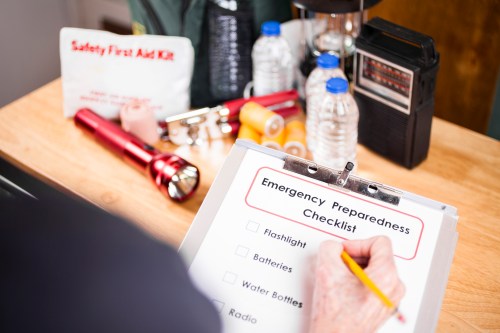 A person writes on a clipboard labeled "Emergency Preparedness Checklist" with supplies like flashlights and water bottles nearby.
