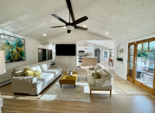 Bright, modern living room with a high ceiling, white walls, a large sofa, yellow ottoman, and open kitchen in the background.