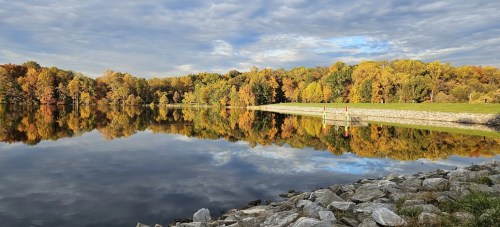 A serene lake reflects colorful autumn trees and a cloudy sky, with a rocky shore in the foreground.