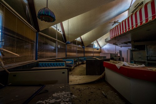 Abandoned diner interior with blue booths, debris on the floor, and a sloped ceiling, illuminated by natural light.
