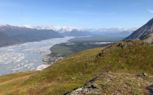 A panoramic view of a river winding through a valley, surrounded by mountains under a clear blue sky.