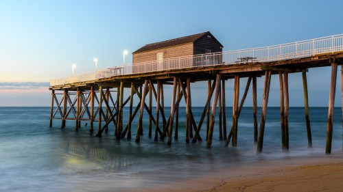 A wooden pier extends over calm waters, featuring a small building at the end, illuminated by soft lights at dusk.