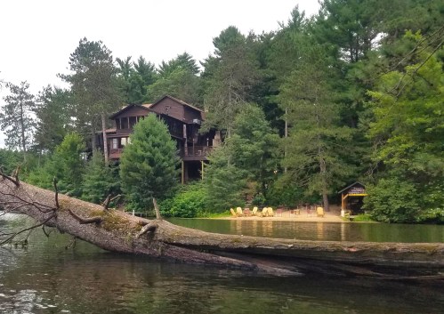 A rustic cabin surrounded by trees, overlooking a calm lake with a fallen log and yellow chairs on the shore.