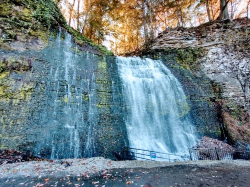 A cascading waterfall flows down a rocky cliff, surrounded by trees and autumn foliage.