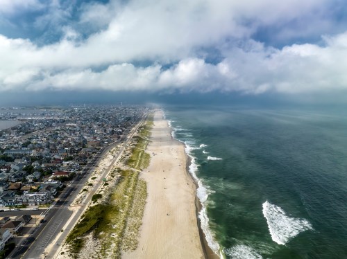 Aerial view of a sandy beach with waves, coastal town, and cloudy sky in the background.