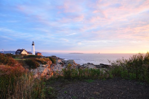 A lighthouse stands by the rocky shore at sunset, with colorful skies and calm waters in the background.