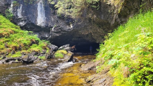 A person stands at the entrance of a dark cave, surrounded by lush greenery and a flowing stream.