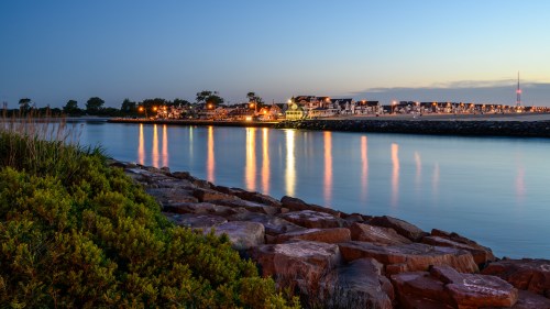 A serene waterfront scene at dusk, with reflections of lights on calm water and rocky shoreline in the foreground.