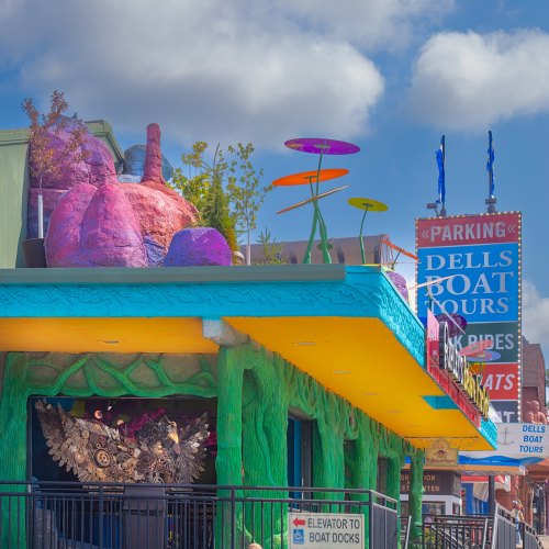 Colorful building with whimsical decor, featuring vibrant plants and a sign for Dells Boat Tours. Bright blue sky above.