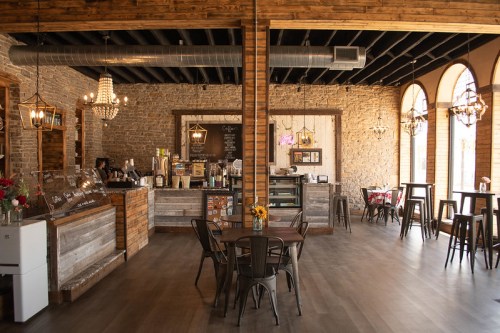 Cozy café interior with wooden beams, stone walls, and a counter displaying coffee and pastries. Sunlight through arched windows.