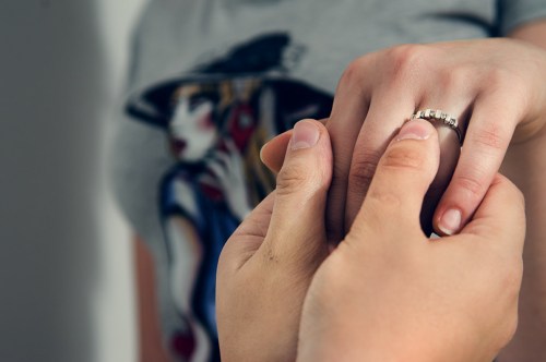 A close-up of two hands, one holding the other, showcasing a ring on the hand being held. A graphic t-shirt is in the background.