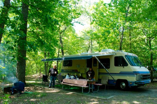A camper parked in a wooded area, with two people cooking and enjoying a meal outside.