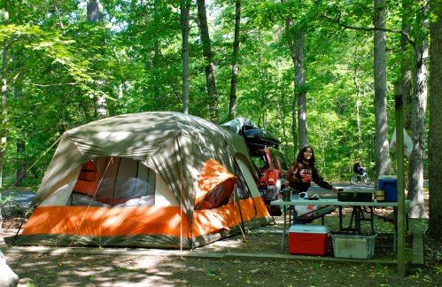 A campsite with a large tent, a woman cooking at a table, and a car parked nearby, surrounded by trees.
