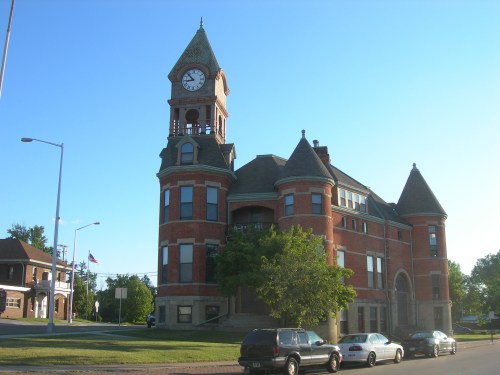 Historic brick building with a clock tower, surrounded by trees and parked cars, under a clear blue sky.