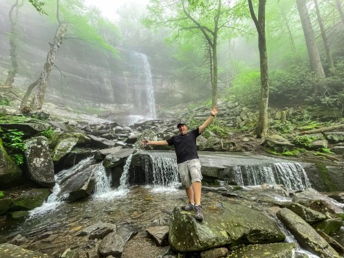 A person stands on rocks by a waterfall in a misty forest, arms raised in excitement amidst lush greenery.