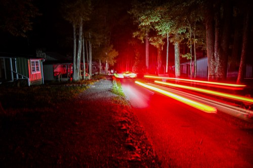 A dark forest road at night, with red light trails from passing cars and illuminated cabins in the background.
