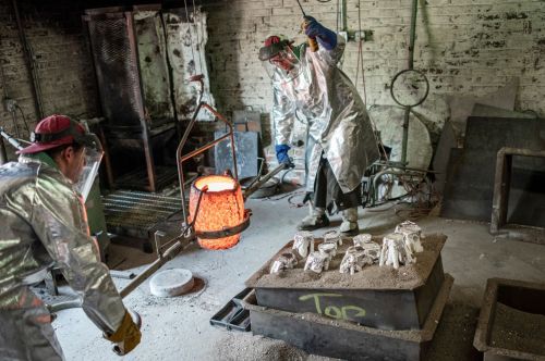 Two workers in protective gear pour molten metal into molds in a workshop, surrounded by tools and equipment.