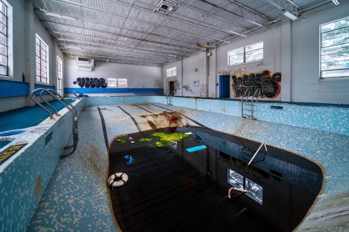 Abandoned indoor swimming pool with dark water, debris, and peeling tiles, surrounded by graffiti on the walls.