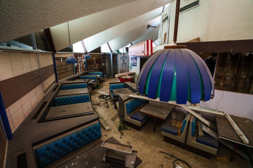 Abandoned restaurant interior with blue booths, broken furniture, and a large pendant light hanging from the ceiling.