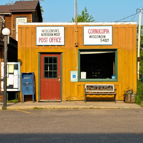 A small wooden post office building with a red door, signs, and a bench outside in Cornucopia, Wisconsin.